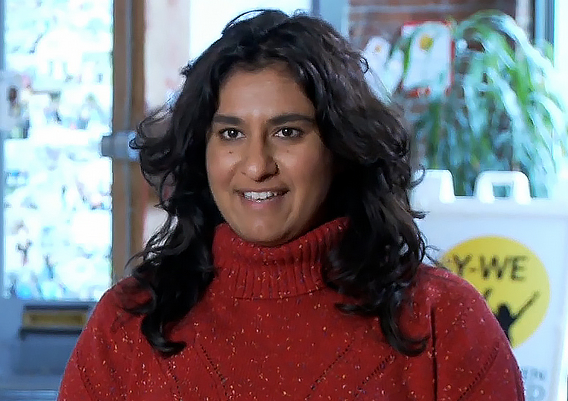 A woman with long dark hair and a red turtle-neck sits for an interview. A sandwich board can be seen in the background that says "Y-WE."