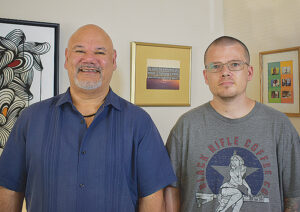 Portrait of a man in a blue shirt, smiling, with a younger man in a grey shirt and glasses to his right.