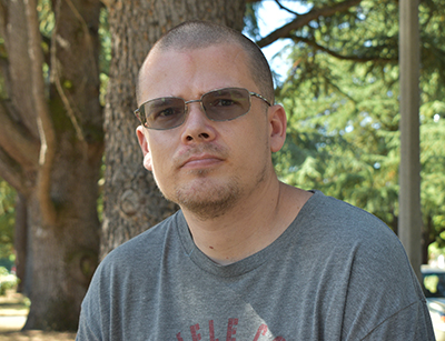 Portrait of a young man in a gray shirt and dark glasses in front of a stand of trees.