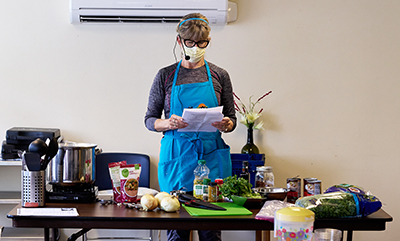 A woman wearing a microphone headset and blue apron stands over a table with onions, a cutting board, and other cooking equipment.