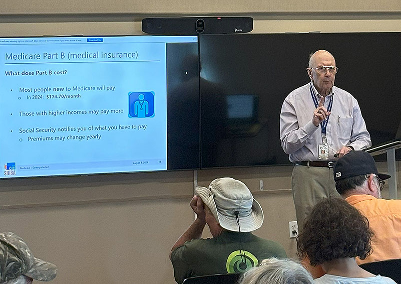 An elderly gentleman wearing a grey button-down shirt, khaki pants, wire-rimmed glasses, and a blue lanyard with name badge speaks at a podium in front of a group of mostly grey-haired people. On the screen behind him is a slide with Medicare information on it.