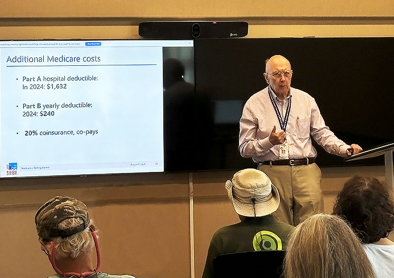 An elderly gentleman wearing a grey button-down shirt, khaki pants, wire-rimmed glasses, and a blue lanyard with name badge speaks at a podium in front of a group of mostly grey-haired people. On the screen behind him is a slide with Medicare information on it.