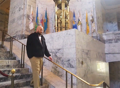 A man in a dark jacket and khakis walks down a staircase below a display of flags on a large marble ledge.