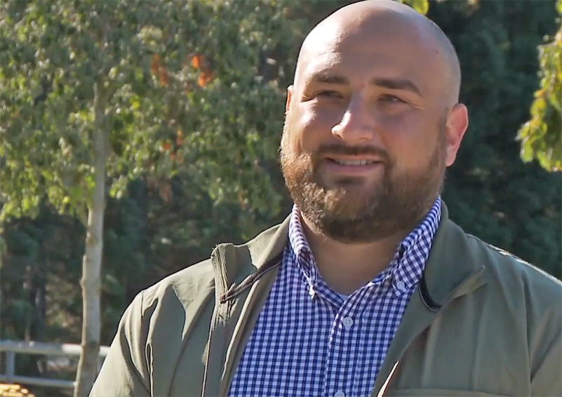 A still image from a video interview with a man with a beard wearing a army green jacket over a blue colored shirts. Trees can be seen out of focus in the background.