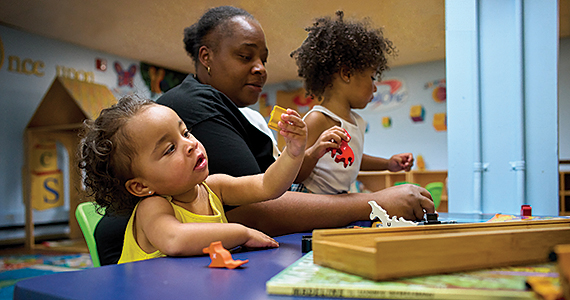 A mom with two toddlers play with dinosaur toys and books at a blue table.