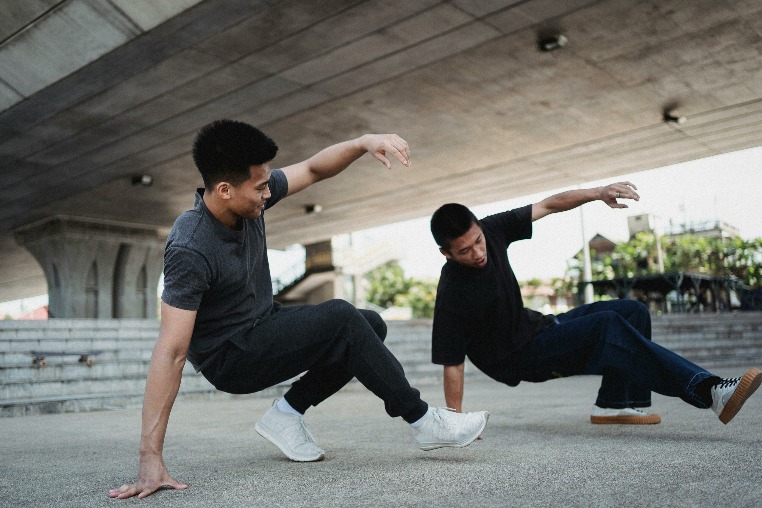 Two young hip hop dancers in matching dark gray and black clothes practice choreography under a bridge.