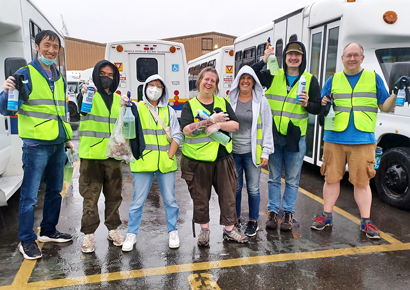 A group of people in neon-yellow vests stand between two buses in a parking lot while holding up spray bottles and other cleaning equipment.