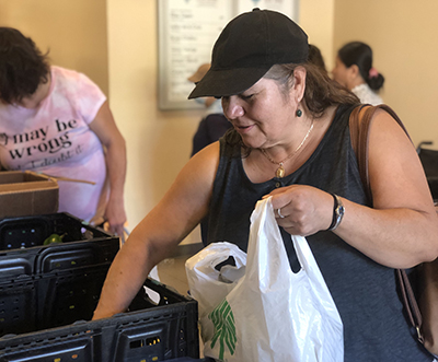 A woman in a black baseball hat reaches into a black plastic bin with one hand while holding a white grocery bag with the other.