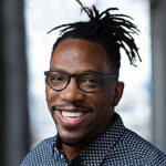 Headshot of a smiling man with his hair in dreads pulled back, glasses, and a blue polka dot shirt. He stands in front of a window.