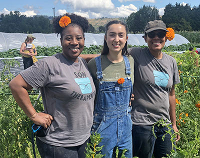 Three women with orange flowers tucked in their hair and clothes stand with arms around in each other in the middle of a farm.