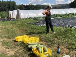 A woman in overalls stands near plastic bins filled with green and yellow squash.