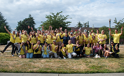 A large group of people in yellow tshirts gesture wildly with their hands while taking a group photo on a grass field.