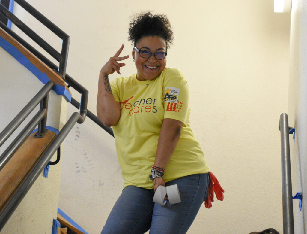 A woman in a yellow t-shirt smiles and makes a peace sign with her fingers while standing in a freshly painted stairwell