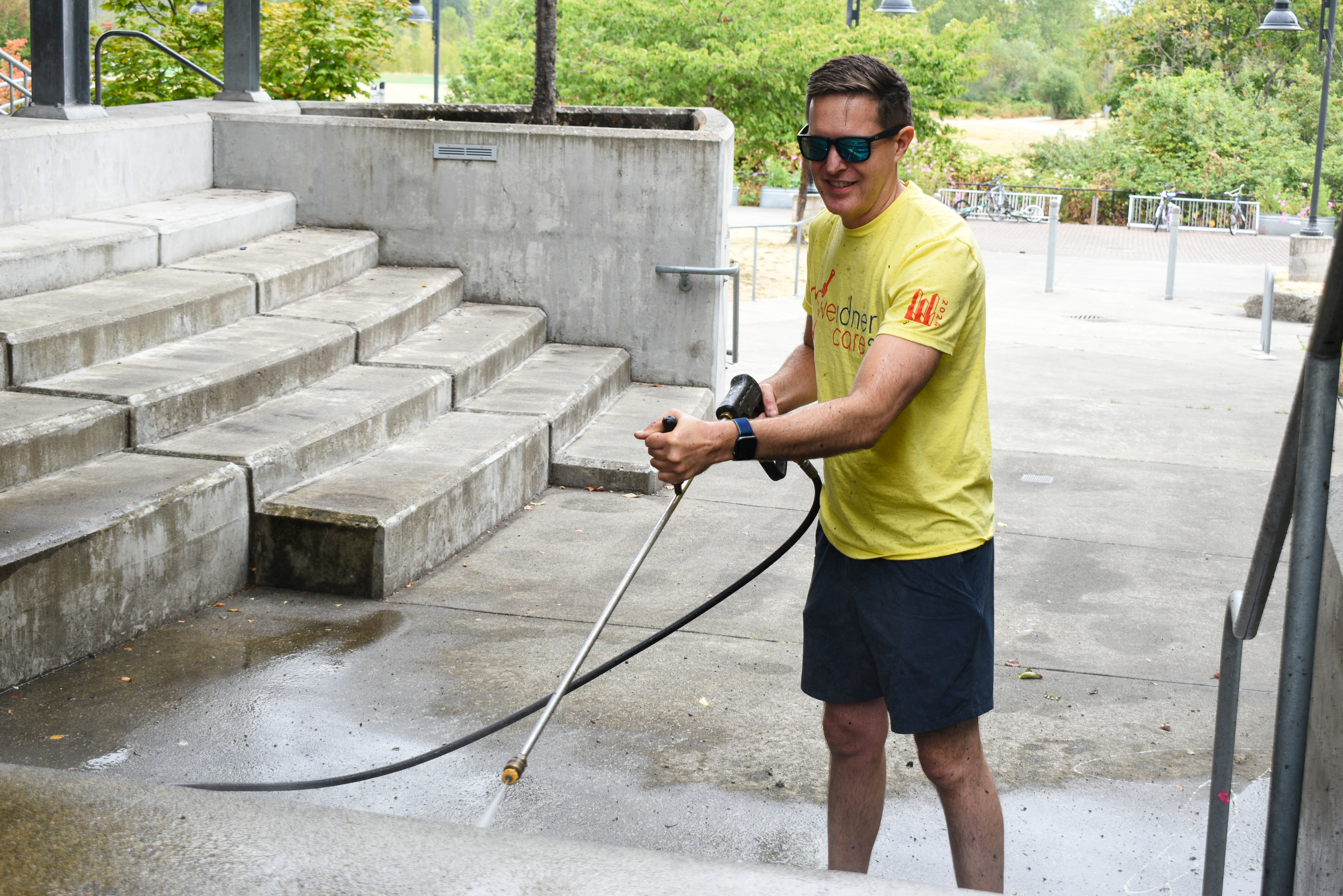 A man in a yellow shirt and shorts cleans a cement staircase with a pressure washer.