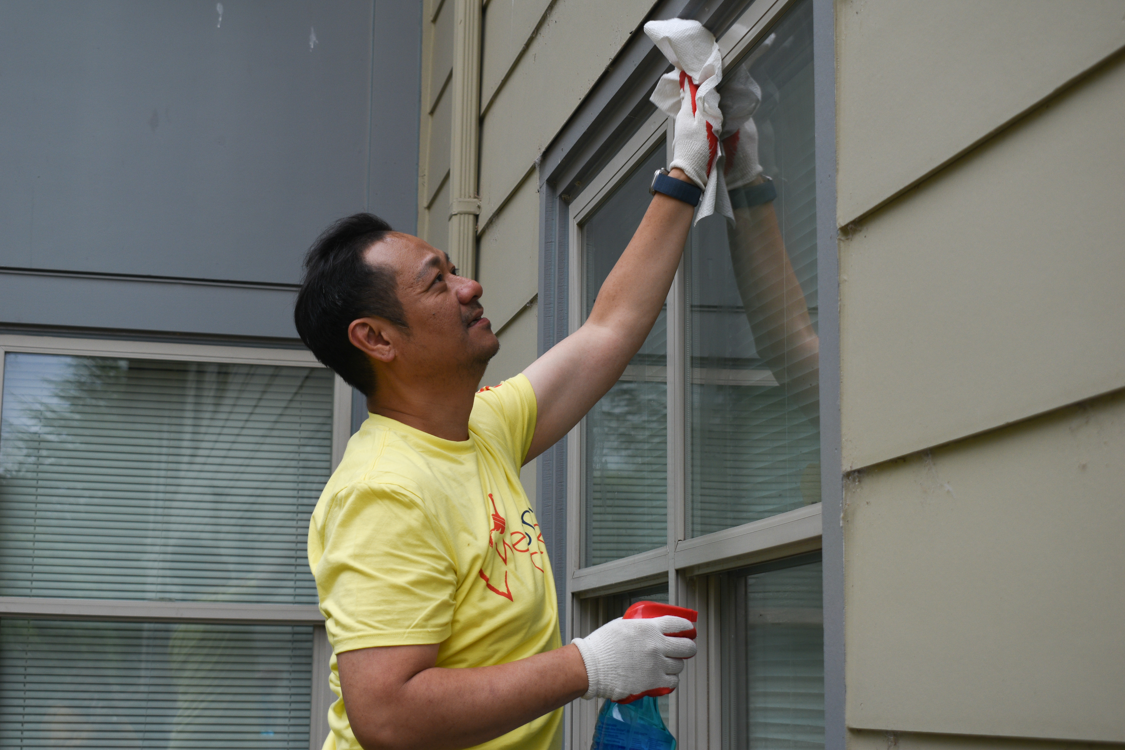 A man in a yellow shirt wipes down the windows of an apartment building after spraying them with glass cleaner.