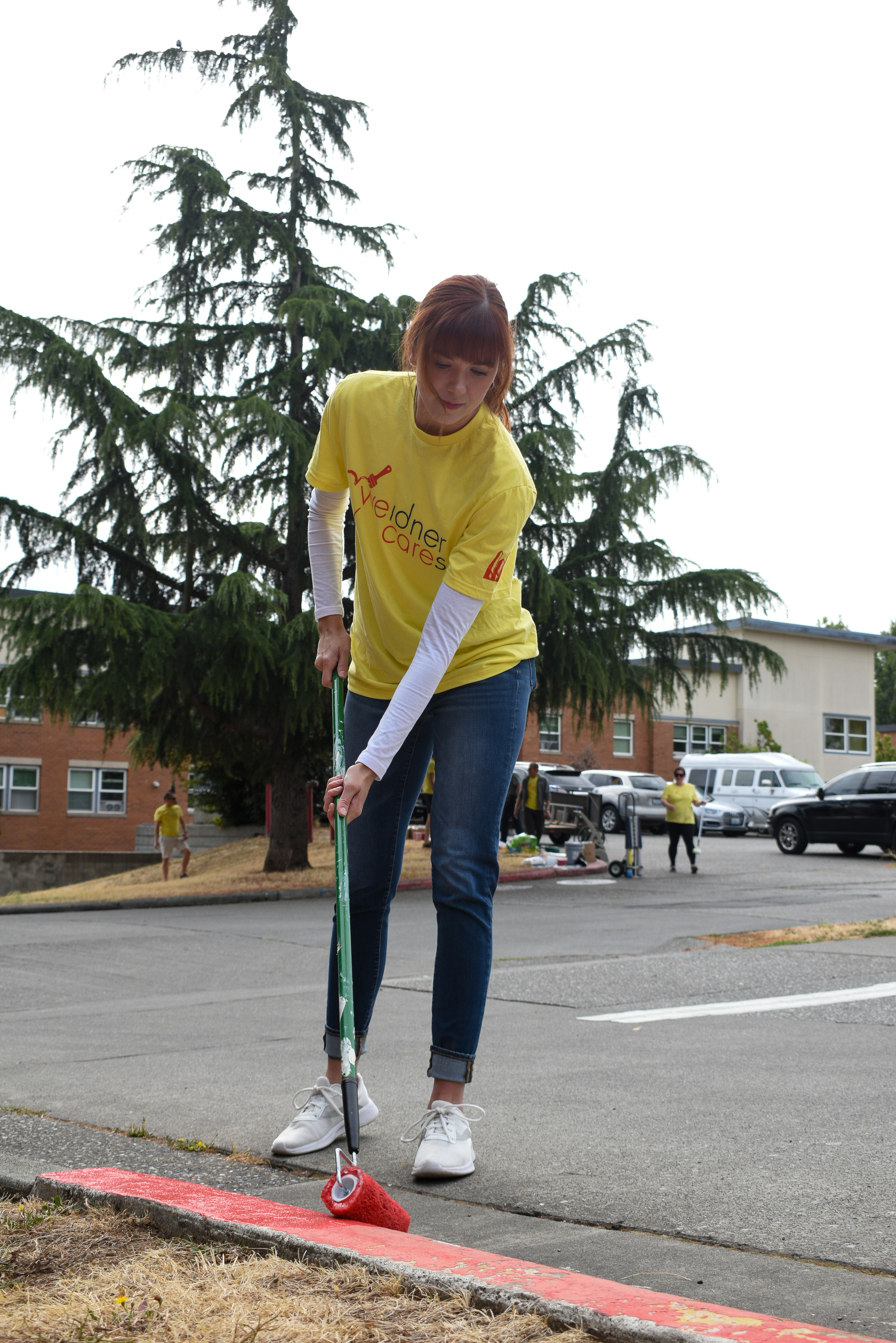 A woman in a yellow shirt uses a paint roller on a pole to apply fresh paint to red fire lane stripping.