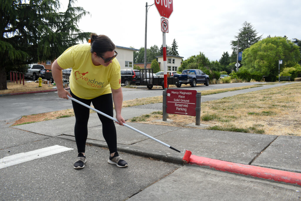 A woman in a yellow shirt uses a paint roller on a pole to apply fresh paint to red fire lane stripping.