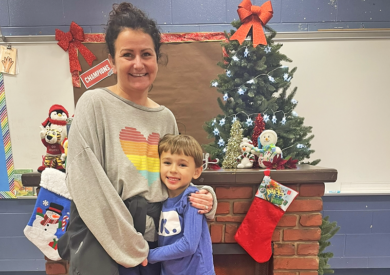 A woman in a gray sweatshirt with a rainbow heart on the front hugs a young boy in a blue sweater in front of a brick fireplace mantel decorated with Christmas stockings and a small tree.