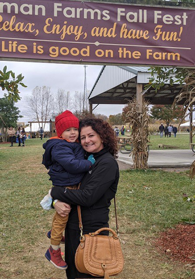 A woman with curly brown hair holds a small child wearing a red knit hat and blue winter coat under a brown banner advertising a fall farm fest with the phrase "Life is good on the farm."