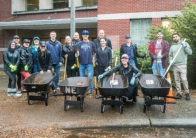 A group of 15 people, most of them in raincoats, pose for a group photo while holding rakes and pitchforks in front of a row of wheelbarrows.