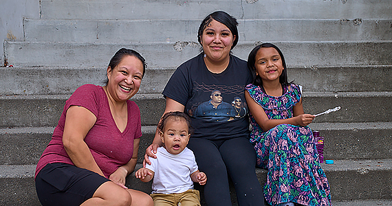 A multigenerational family sit on concrete steps, smiling. They include a grandmother, mother, young daughter, and baby boy.