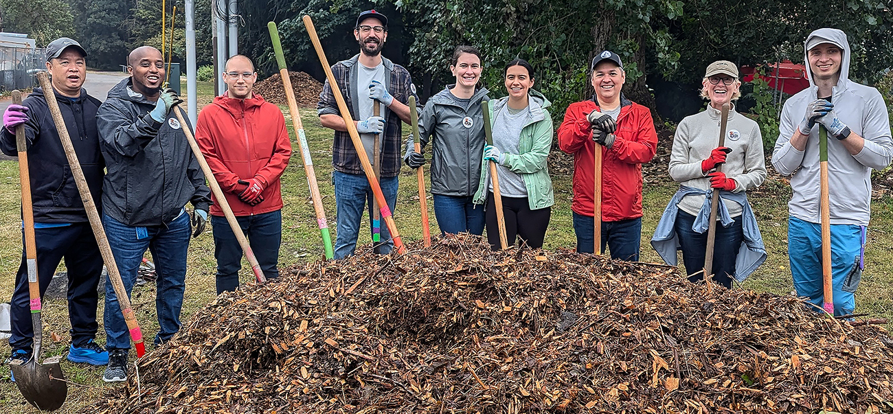 A smiling group of 9 people dressed for cool weather and holding gardening shovels pose in a semicircle behind a large pile of mulch.