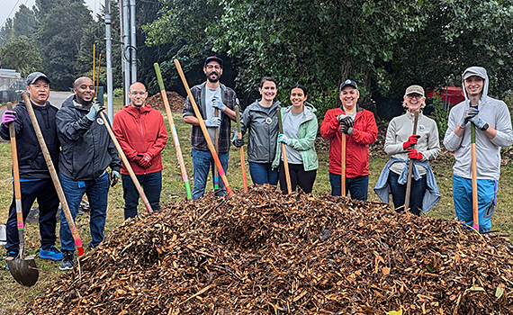 A smiling group of 9 people dressed for cool weather and holding gardening shovels pose in a semicircle behind a large pile of mulch.