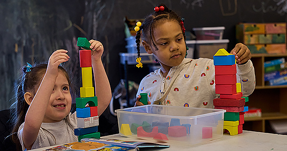 Two young girls play with colorful blocks on a white table. There's a book open in front of them, and behind them is a chalkboard.