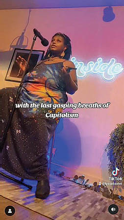 Screenshot from a Tik Tok video of a woman in hazy purple, neon light reciting poetry into a microphone. Captioning in white text reads: 'with the last gasping breaths of Capitalism.'