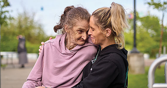 An elderly woman with light-brown hair in a ponytail and wearing a pink hoodie, smiles and touches foreheads with a middle-aged woman with blonde hair in a ponytail and wearing a black hoodie.