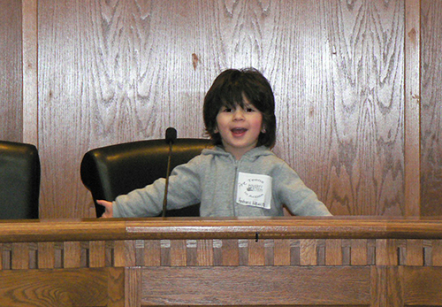 A little boy with long dark hair and a gray sweatshirt smiles big and extends his arms out wide behind a wooden podium with a microphone.