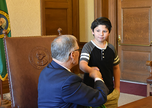 A man with thin gray hair and blue suit reaches beside him to shake the hands of a young boy.