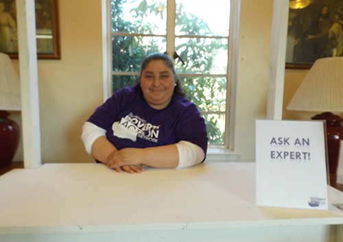 A woman in a purple t-shirt that reads "Statewide Poverty Action Network" sits behind a white desk with a sign that says "Ask the expert."