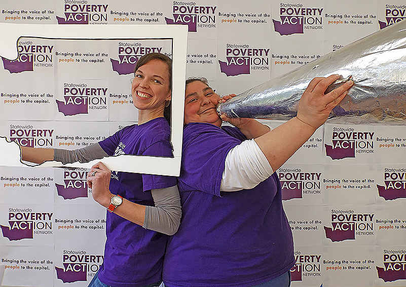 Two women in bright purple T-shirts pose back-to-back in front of a a photobooth step-and-repeat with purple Statewide Poverty Action Network logos on it. One looks through an empty picture frame in the shape of Washington state; the other holds a giant megaphone.