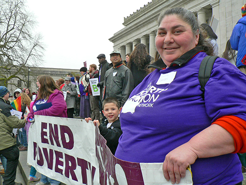 A woman in a purple t-shirt that reads "Statewide Poverty Action Network" and a young boy in a black jacket hold up a banner that says "End Poverty" in front of a crowd of people standing on the steps of a large white building with Greek columns.