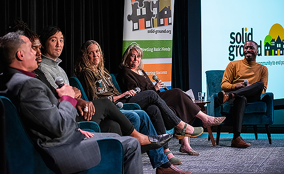 Six people sit in blue armchairs on a stage. The man to the far left speaks into a microphone as the others listen intently. Behind them is a green, orange, and black pullup banner and a screen with the Solid Ground logo on it.