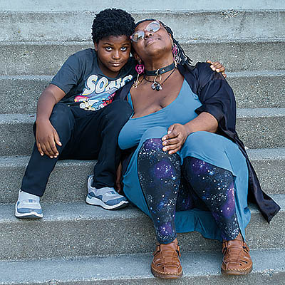 A Black mother and young son sit with arms around each others' shoulders on a set of concrete stairs.