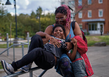 A Black woman with pink braids and glasses hugs her young son as he plays on a jungle gym. Behind them is a street lamp and an apartment building.