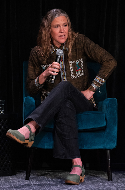 A woman with long wavy blonde and gray hair crosses her legs while speaking into a microphone and sitting on a plush blue chair on a darkly lit stage.