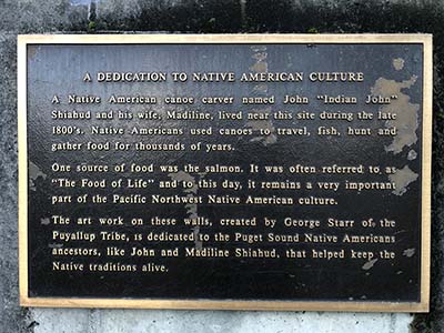 A picture of a plaque reading "A DEDICATION TO NATIVE AMERICAN CULTURE. A Native American canoe carver named John "Indian John" Shiahud and his wife, Madiline, lived near thsi site during the late 1800's. Native Americans used canoes to travel,fish, hunt and gather food for thousands of years. One source of food was the salmon. It was often referred to as "The Food of Life" and to this day, it remains a very important part of the Pacific North West Native American Culture. The art work on theses walls, created by George Starr of the Puyallup Trible, is dedicated to the Puget Sound Native American ancestors, like John and Madiline Shiahud, that helped keep the Native traditions alive."