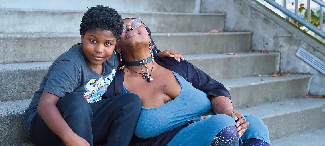 A young Black boy and his mom sit on concrete steps. His arm is thrown around her shoulder and he smiles into the camera. Her eyes are closed, and she leans back onto his shoulder.