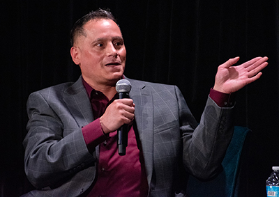 A man in a gray sports coat over a maroon dress shirt gestures with one hand while speaking into a microphone held in the other.