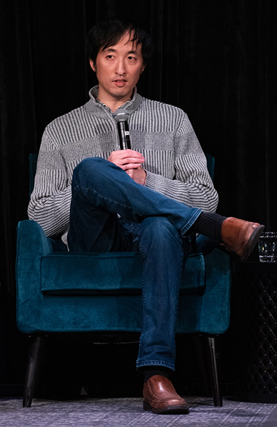 A man in a gray sweater and jeans speaks into a microphone and sitting on a plush blue chair on a darkly lit stage.