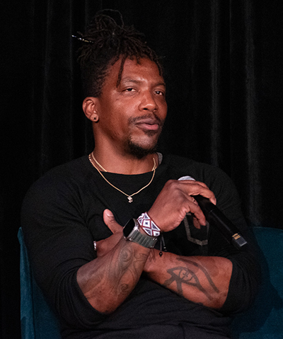 A man with braided hair and a black t-shirt cross his tattooed arms as he talks into microphone.