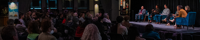 A wide shot of a group of people in blue plush chairs on a stage addressing a darkened room full of people.