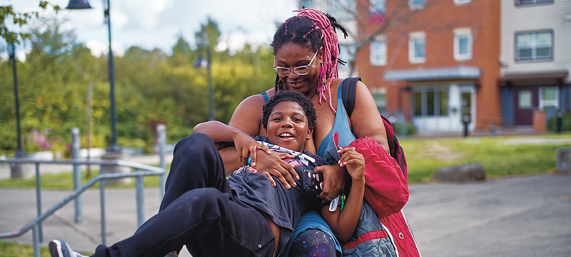 A Black woman with pink braids and glasses hugs her young son as he plays on a jungle gym. Behind them is a street lamp and an apartment building.