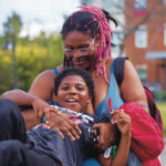 A Black woman with pink braids and glasses hugs her young son as he plays on a jungle gym. Behind them is a street lamp and an apartment building.