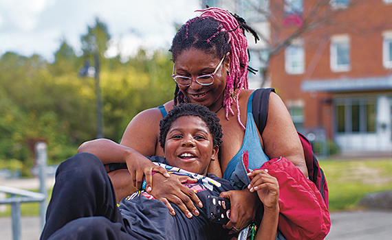 A Black woman with pink braids and glasses hugs her young son as he plays on a jungle gym. Behind them is a street lamp and an apartment building.