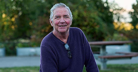 A grey-haired man wearing a navy-blue pullover smiles broadly for the camera. He sits outdoors on a bench.