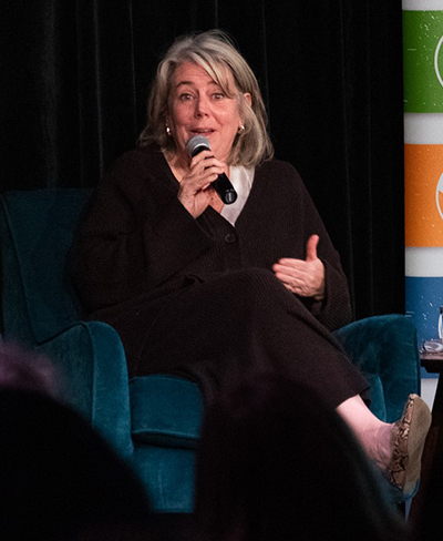 A woman with blonde and gray hair gestures while speaking into a microphone and sitting on a plush blue chair on a darkly lit stage.